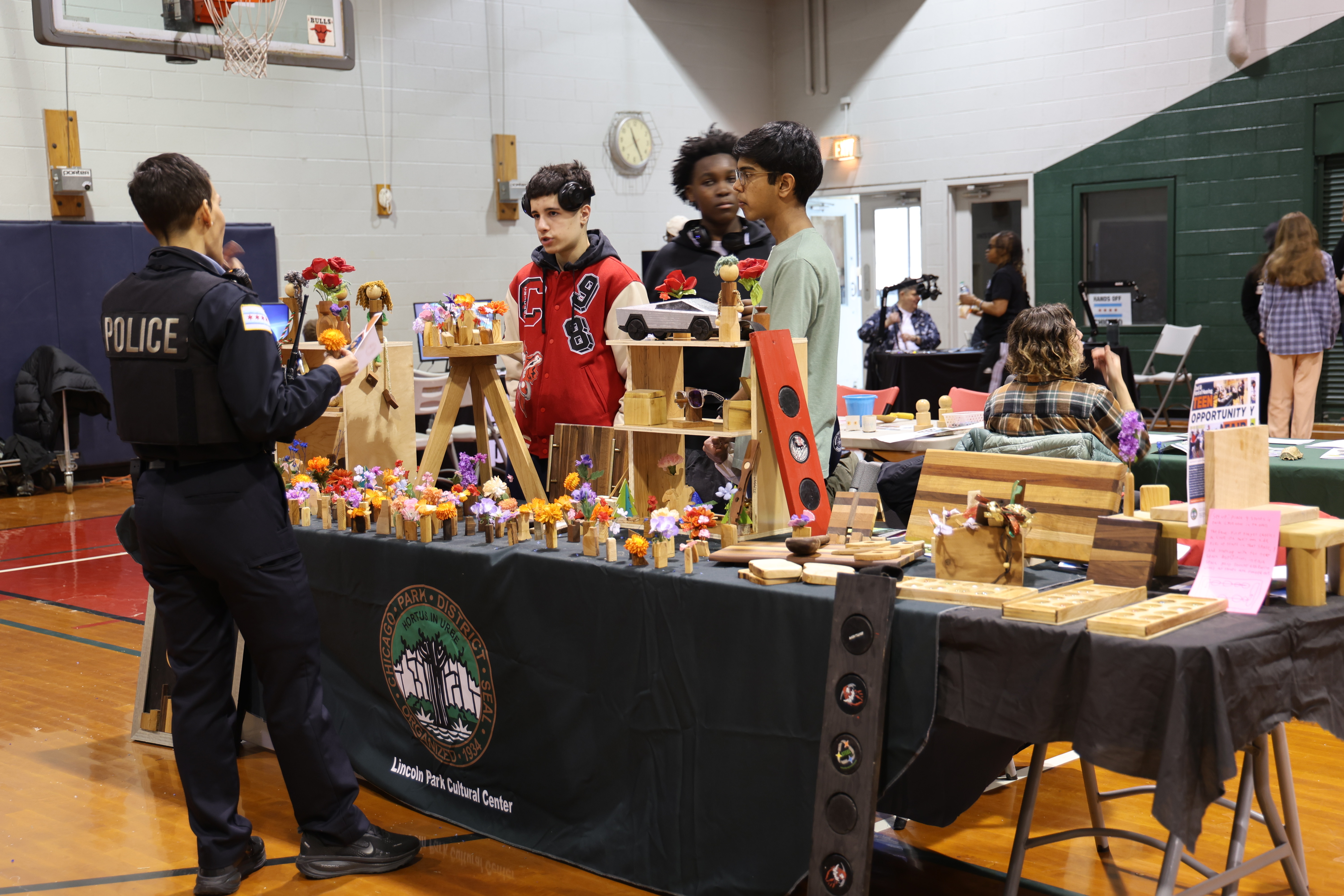 Police officer talks to three teens at a craft fair table with flowers in a gym.